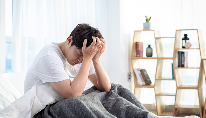 남성 기능 변화 안내에 사용된, 침대에 앉아 두 손으로 머리를 감싼 남성 사진