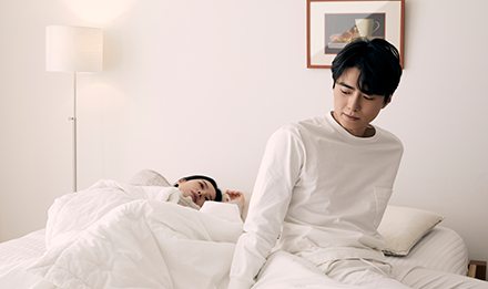 침대에 앉은 남성이 옆에 누운 여성을 돌아보는 모습